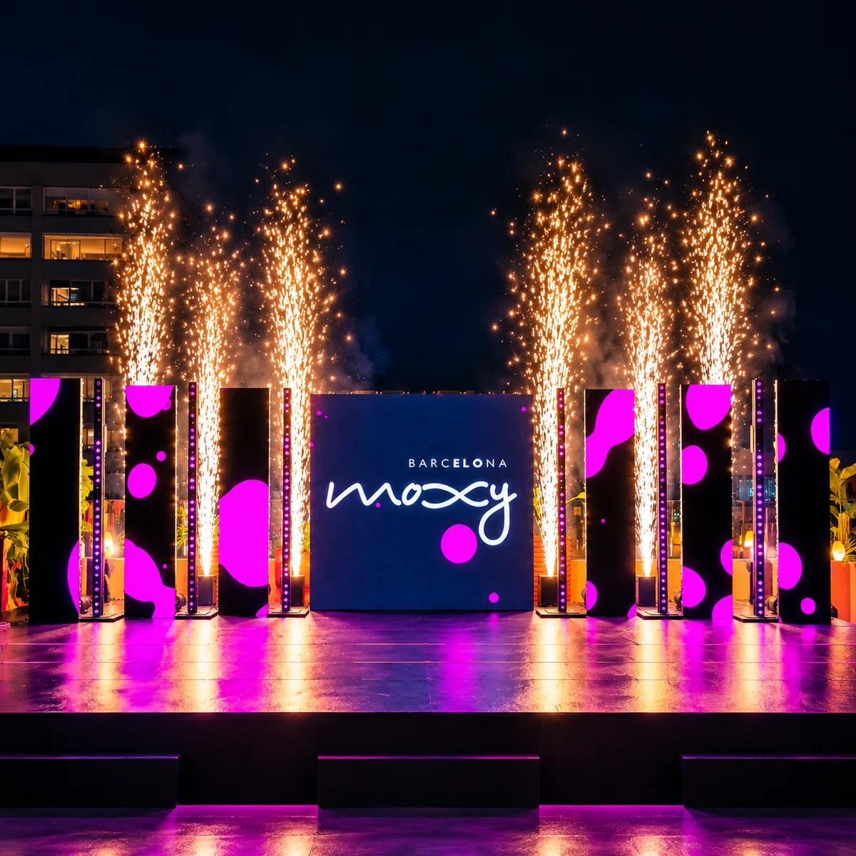 Escenario nocturno del aniversario de Moxy Barcelona, con surtidores de chispas y tótems de LED con lunares rosas.